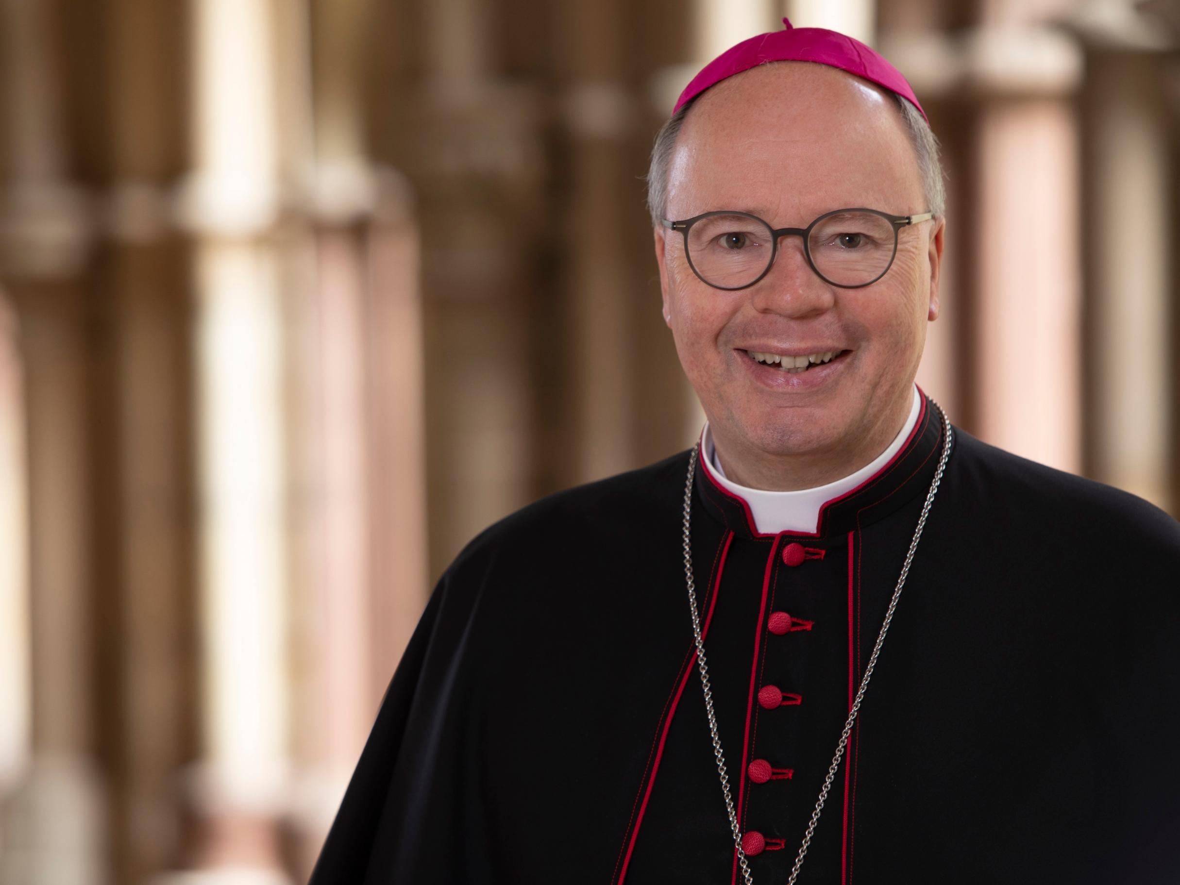 Hier ist ein Portrait von Bischof Ackermann, lächelnd im Kreuzgang zu sehen. Er Trägt ein Ornat.